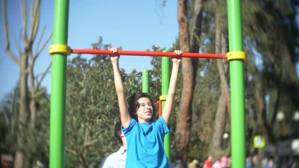 Een knappe man in lichte kleren leert een jongen in een blauw T-shirt zelf optrekken correct op een horizontale balk in een zonnige zomer park, 4k, slow-motion — Stockvideo