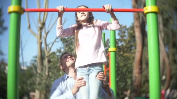 A handsome man teaches a girl in a pink T-shirt how to pull himself up correctly on a horizontal bar in a summer sunny park, 4k, slow motion — Stock Video