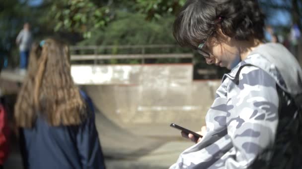 Een jongen die alleen met behulp van een mobiele telefoon in de buitenlucht. terwijl andere kinderen actieve rust hebben. Smartphone verslaving. 4k, slow-motion — Stockvideo