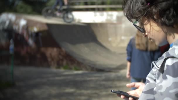 Um rapaz sozinho a usar um telemóvel ao ar livre. enquanto outras crianças têm descanso ativo. Vício em smartphones. 4k, câmera lenta — Vídeo de Stock
