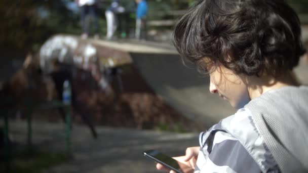 Un niño solo usando un teléfono móvil al aire libre. mientras otros niños descansan activamente. Adicción a teléfonos inteligentes. 4k, cámara lenta — Vídeo de stock