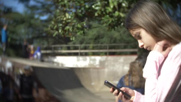 A lány egyedül használatával egy mobil telefon a szabadban. míg más gyerekek állanak. Smartphone függőség. 4k, lassú mozgás — Stock videók