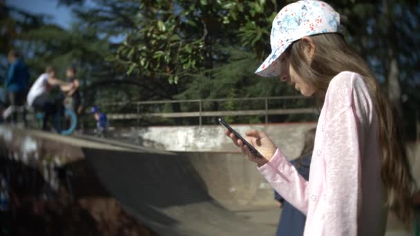 Une fille seule utilisant un téléphone portable à l'extérieur. tandis que les autres enfants ont un repos actif. Dépendance au smartphone. 4k, au ralenti — Video