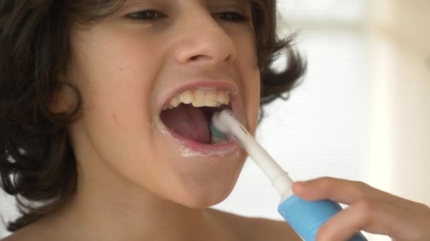 Ребенок чистит зубы перед зеркалом в 4k. Мальчик-подросток чистит зубы электрической зубной щеткой, крупным планом, снимает замедленную съемку — стоковое видео