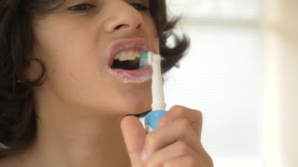Barnet rengör tänderna framför en spegel i 4 k. pojke tonåringen borstar tänderna med eltandborste, närbild, Slowmotion-skytte — Stockvideo