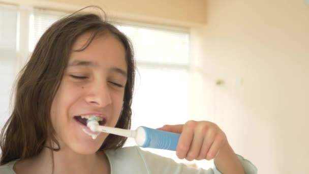 Ребенок чистит зубы перед зеркалом в 4k. Девушка-подросток чистит зубы электрической зубной щеткой, крупным планом, снимает замедленную съемку — стоковое видео