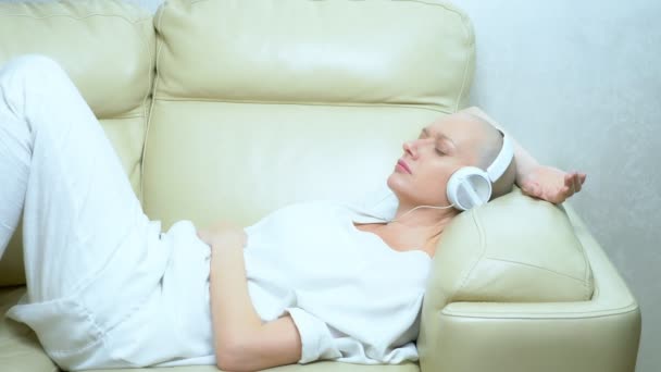 Hermosa chica calva en los auriculares disfruta de la música en casa en el sofá — Vídeo de stock