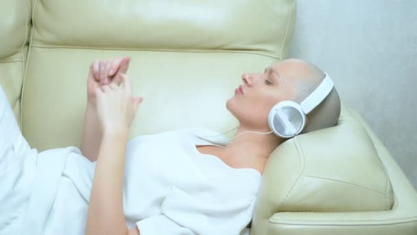 Όμορφη φαλακρή κοπέλα με ακουστικά απολαμβάνει τη μουσική στο σπίτι στον καναπέ — Αρχείο Βίντεο