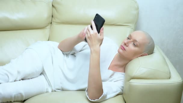 Bela mulher careca usa um smartphone enquanto deitado em um sofá na sala de estar — Vídeo de Stock