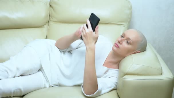 Beautiful bald woman uses a smartphone while lying on a sofa in the living room — Stock Video