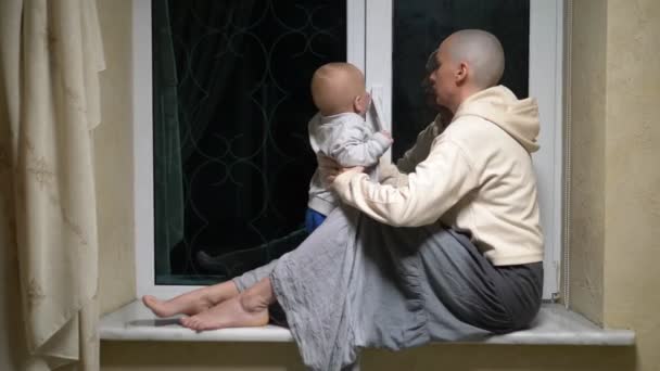 Skallig kvinna och barn sitta tillsammans vid fönstret på kvällen. — Stockvideo