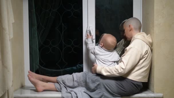 ハゲの女性と赤ちゃんが夜の窓の前に座り. — ストック動画