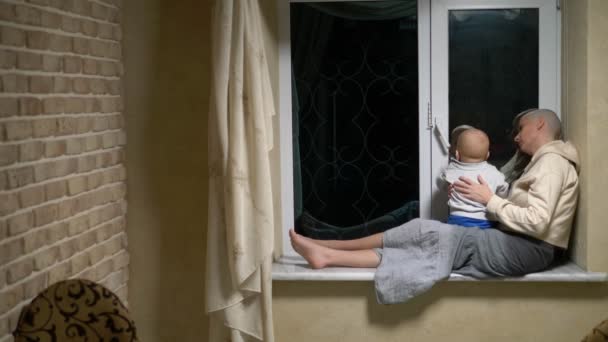 Frau mit Glatze und Baby sitzen abends zusammen am Fenster. — Stockvideo