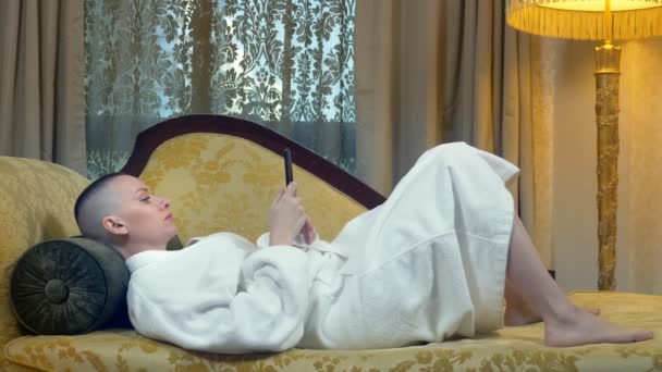 Красивая лысая женщина в халате лежит на диване с телефоном вечером на заднем плане. окна в роскошном интерьере — стоковое видео