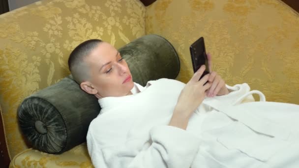 Όμορφη φαλακρή γυναίκα με μπουρνούζι ξαπλώνει σε καναπέ με τηλέφωνο το βράδυ στο βάθος. ενός παραθύρου σε ένα πολυτελές εσωτερικό — Αρχείο Βίντεο