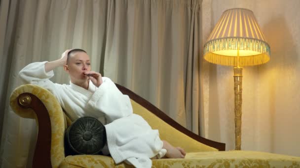 Uma linda garota careca em um roupão de banho fica em um sofá luxuoso no fundo de uma janela e fuma um cigarro eletrônico. espaço de cópia — Vídeo de Stock