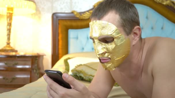 Hombre guapo en una máscara de oro se relaja en una cama de lujo con un teléfono celular. primer plano — Vídeos de Stock