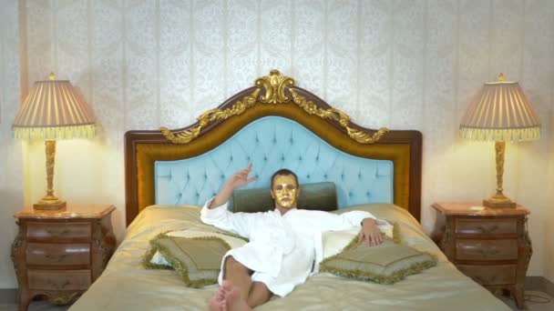 Un hombre guapo con una máscara dorada y un albornoz descansa sobre una cama de lujo. mirando a la cámara — Vídeo de stock
