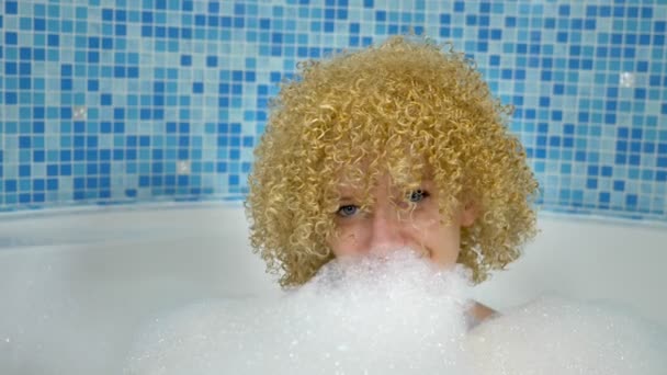 Joyful caucasian blond woman enjoys a home spa in the bathroom, lies in a foam bath with a white mask on her face, plays with foam clouds — Stock Video