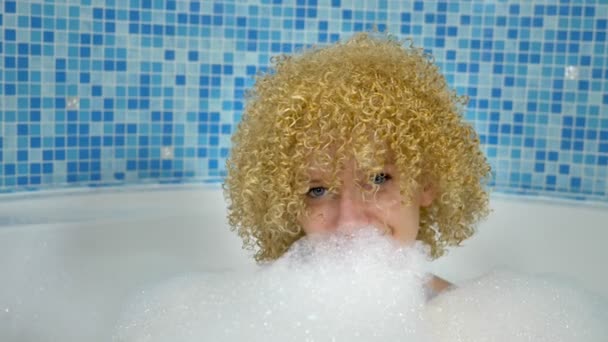 Alegre mujer rubia caucásica disfruta de un spa en el baño, se encuentra en un baño de espuma con una máscara blanca en la cara, juega con nubes de espuma — Vídeos de Stock