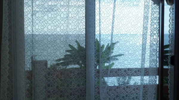 Przezroczysta zasłona na oknie z widokiem na morze, delikatnie poruszona przez wiatr. Promienie słońca. — Zdjęcie stockowe