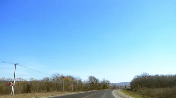 Vista desde el parabrisas de un coche. En un día soleado, un camino vacío corre entre campos y arbustos en el otoño. cielo azul claro. un giro es visible delante — Foto de Stock