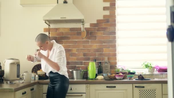 Medelstora skott. Snygg skallig kvinna dricker en varm drink när du står i köket hemma. — Stockvideo