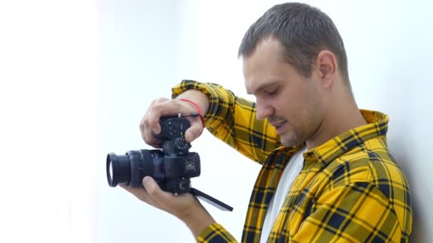 Joven guapo con una camisa amarilla a cuadros, fotógrafo profesional sosteniendo una cámara toma fotos. Fotógrafo en el trabajo . — Vídeos de Stock