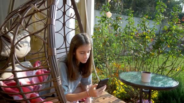 Linda chica adolescente descansando en una silla columpio en el patio trasero usando un teléfono inteligente — Vídeos de Stock