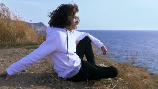 Teen boy sitting on the edge of a cliff over the sea. the hair at his curly hair — Stockvideo