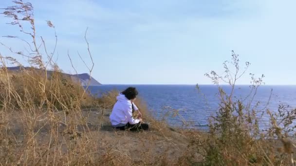 Adolescente sentado en el borde de un acantilado sobre el mar. el pelo en su pelo rizado — Vídeo de stock