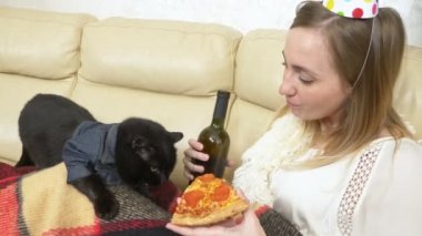 Parti şapkalı yalnız bir kız, şişeden şarap içiyor ve kanepede kedisiyle pizza yiyor.
