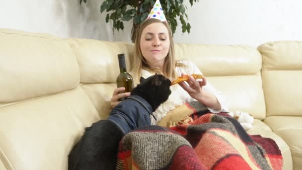 Een eenzaam meisje met een feestmuts, wijn drinkend uit de fles en pizza etend met een kat op de bank — Stockvideo