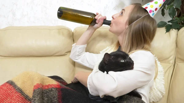 Una ragazza sola con un cappello da festa, che beve vino dalla bottiglia e mangia pizza con un gatto sul divano — Foto Stock