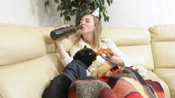Una ragazza sola con un cappello da festa, che beve vino dalla bottiglia e mangia pizza con un gatto sul divano — Foto Stock