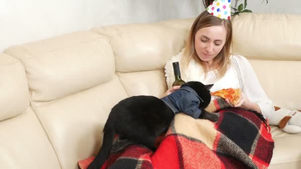 Una ragazza sola con un cappello da festa, che beve vino dalla bottiglia con un gatto — Video Stock