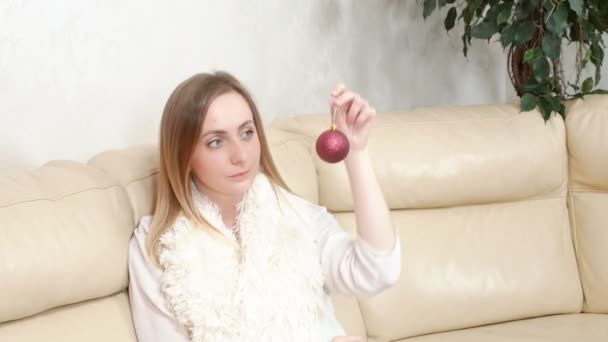 Szőke csodálja a fényes piros karácsonyi labdát, ül a kanapén — Stock videók