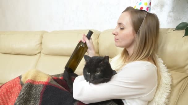 Una chica solitaria en un sombrero de fiesta, bebiendo vino de la botella con un gato — Vídeos de Stock