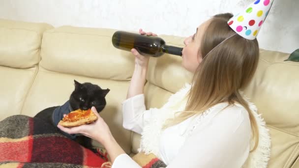 Una chica solitaria en un sombrero de fiesta, bebiendo vino de la botella con un gato — Vídeo de stock