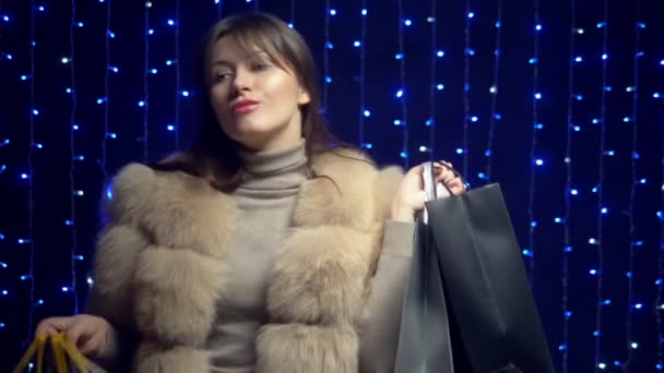 Стильная девушка в меховом пальто с пакетами на фоне гирлянд. продажа товаров — стоковое видео