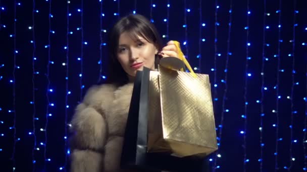 Stylish girl in a fur coat with packages on a background garlands. shopping sale — Stock Video