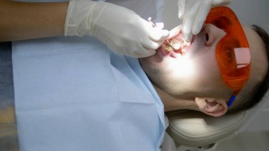 Yaklaştın. Eldivenli bir dişçi bir adamın dişlerini muayene eder.