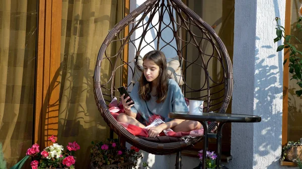 Słodkie nastolatek dziewczyna odpoczywa w bujany krzesło w podwórku za pomocą telefon komórkowy — Zdjęcie stockowe