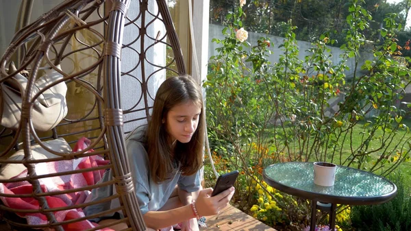 Słodkie nastolatek dziewczyna odpoczywa w bujany krzesło w podwórku za pomocą telefon komórkowy — Zdjęcie stockowe