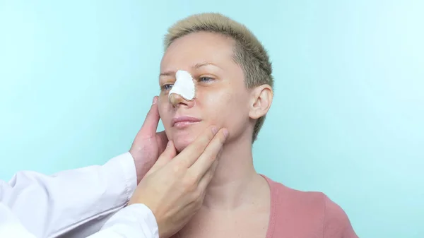 Las manos del médico tocan la cara de una mujer con un vendaje en la nariz . — Foto de Stock