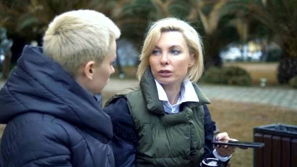 Twee volwassen blonde vrouwen praten, zitten op een bankje in het park. — Stockvideo