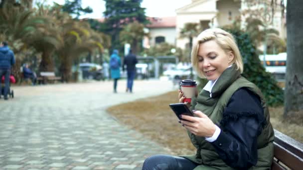 Stijlvolle blonde vrouw zittend op een bankje in het park met koffie en telefoon — Stockvideo