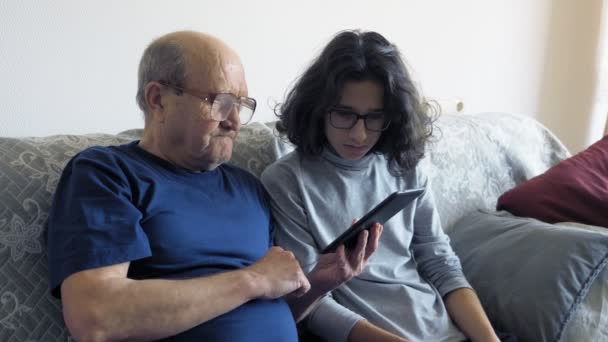 Nagyapa és unokája elektronikus könyvet használnak. ül a kanapén a szobában — Stock videók