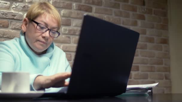 高齢の女性はノートパソコンで作業し、テーブルに座っています。高齢者の仕事のコンセプト — ストック動画