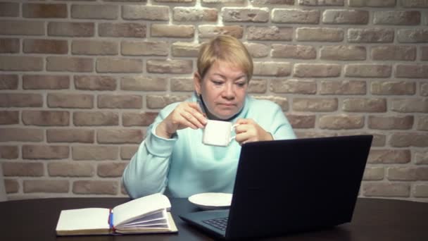 Una anciana trabaja con un portátil, sentada en una mesa. concepto de trabajo de edad avanzada — Vídeos de Stock